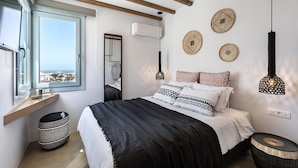 A bedroom with a large window, a bed with patterned bedding, a hanging light fixture, and a wall-mounted air conditioner. - Santo Bloom Luxury Villas with Sea View & Two Pools (Santorini)