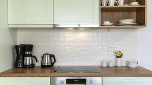 A kitchen with light green cabinets, a stainless steel range hood, a Bosch oven, a coffee maker, and a kettle. There are white tiles on the backsplash and a wooden shelf above the counter with cups and plates. - Santo Bloom Luxury Villas with Sea View & Two Pools (Santorini)