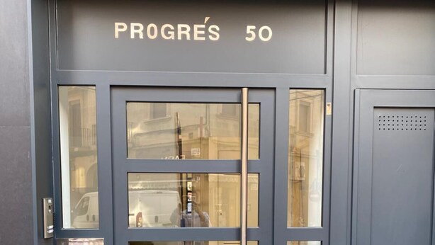 Ein moderner Gebäudeeingang mit einer dunkelblauen Tür, Glasfeldern und der Aufschrift "PROGRÈS 50" darüber.