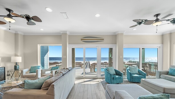 Un vaste salon avec vue sur l’océan, aménagé avec des canapés de couleur beige et turquoise, et doté de deux ventilateurs de plafond.