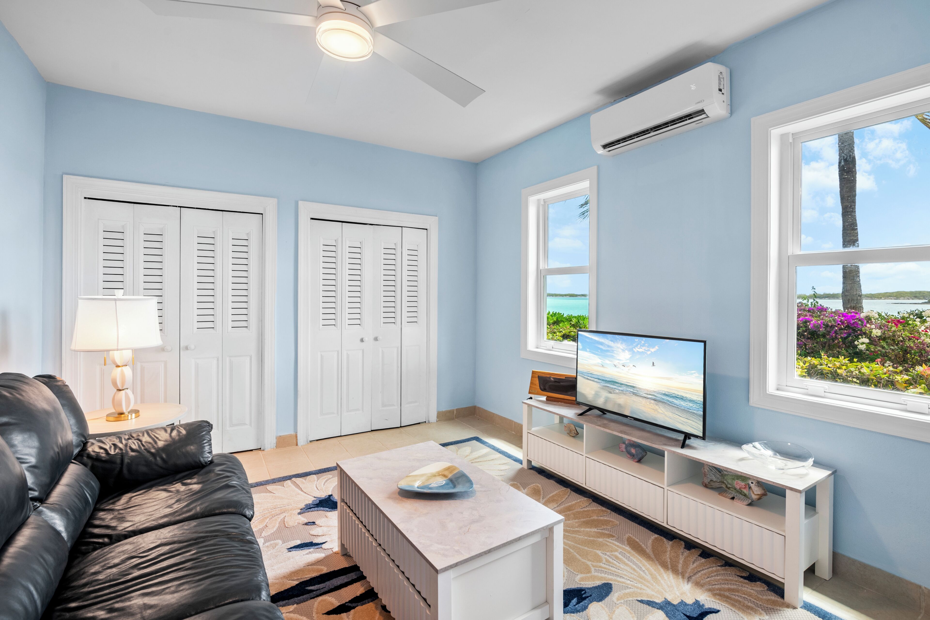 Un salón con televisor, armarios blancos, ventilador de techo y vista al océano a través de una ventana.