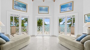 Ein geräumiges Zimmer mit hohen Decken, weißen Wänden und einem Balkon mit Meerblick. Es gibt zwei beigefarbene Sofas mit blauen Kissen, eine Holzbalkendecke und gerahmte Bilder an den Wänden.
