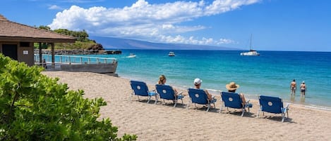 En strand med solstolar och en liten byggnad, med utsikt över ett lugnt hav med båtar.