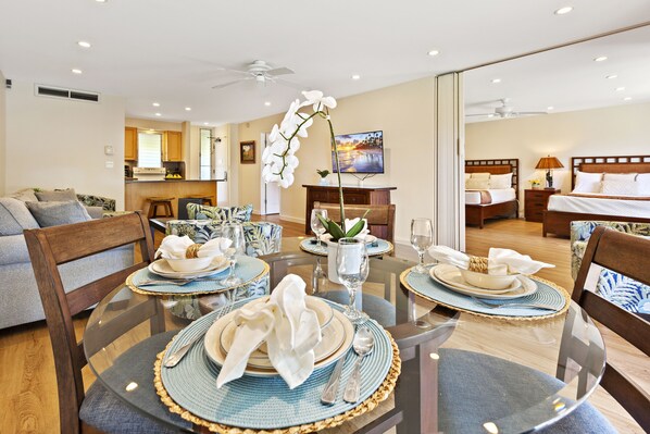 A dining area with round tables, chairs, and a central orchid. A kitchen and a bedroom are visible in the background.