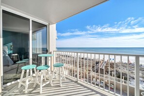 Un balcón con vista a la playa, que incluye un ventanal de vidrio, dos mesitas redondas y sillas blancas.