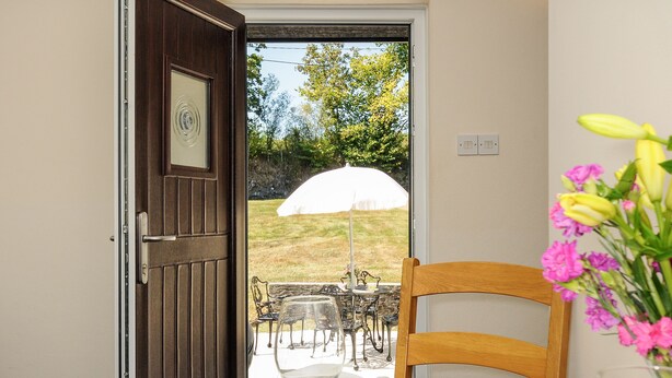 Puerta abierta que da a un área de comedor al aire libre con una mesa dispuesta para dos personas, una silla y un paraguas blanco.