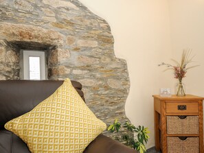 Una pared de piedra, un sofá de cuero marrón con un cojín estampado, una mesita de madera con un jarrón de flores y una planta en maceta.