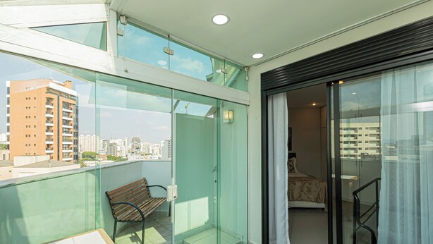 A balcony with a glass door leading to a bedroom, a bench, and a table. - OFP - Duplex na Oscar Freire By Anora Spaces (São Paulo)