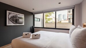 Ett hotellrum med en stor säng, två handdukar med en logotyp, en inramad tavla på väggen och utsikt över byggnader genom fönstret.