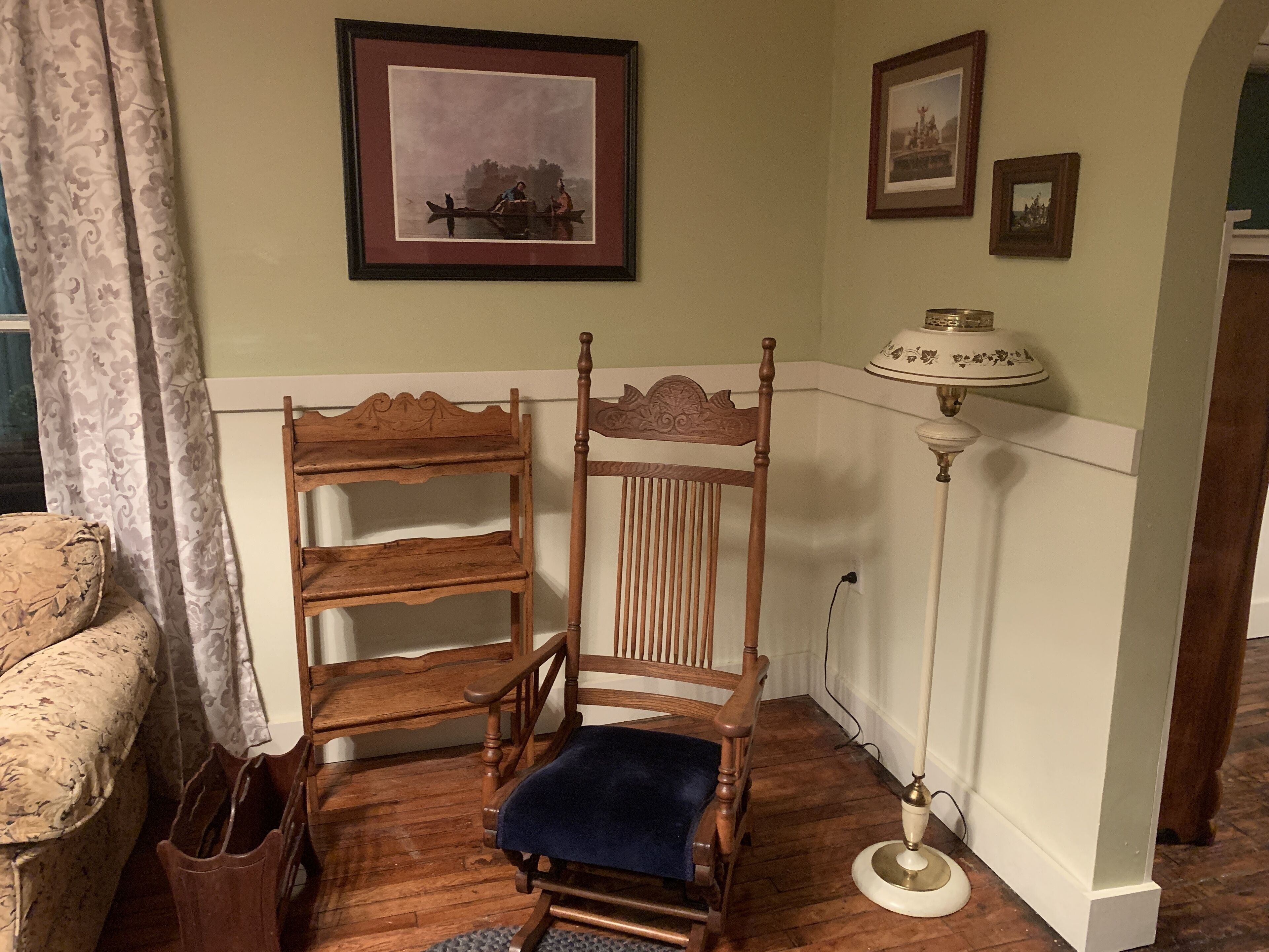 A corner room with a wooden chair, a small wooden shelf, a lamp, and a framed picture on the wall.