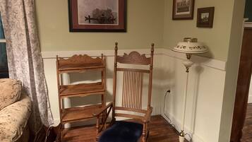 A corner room with a wooden chair, a small wooden shelf, a lamp, and a framed picture on the wall.