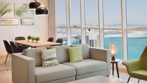 Una sala de estar moderna con un sofá, una mesa de centro y un comedor. Amplias ventanas ofrecen vistas a un paisaje urbano y una masa de agua.