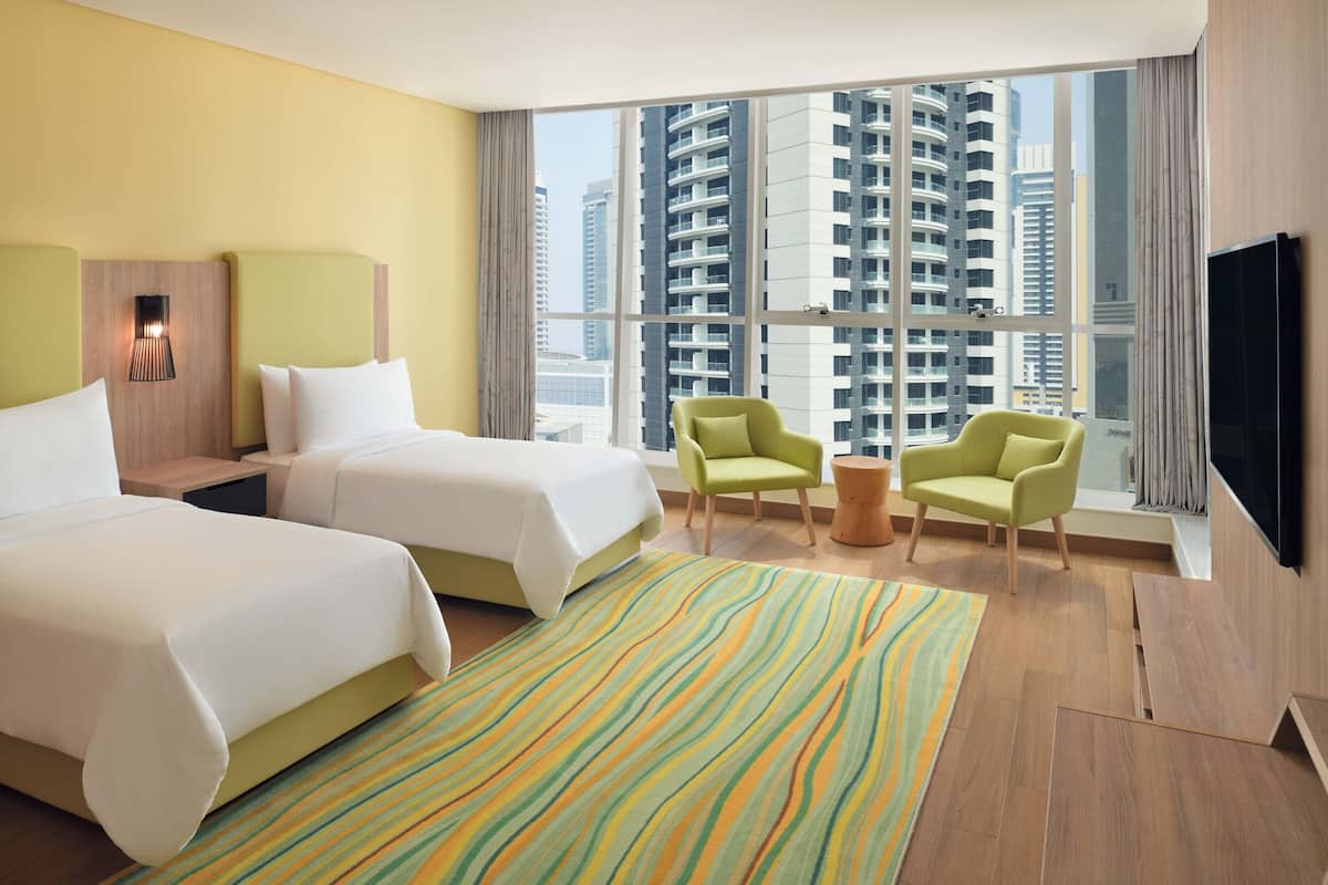 Habitación de hotel con dos camas, televisión, ventana con vista a la ciudad y un pequeño espacio de descanso.