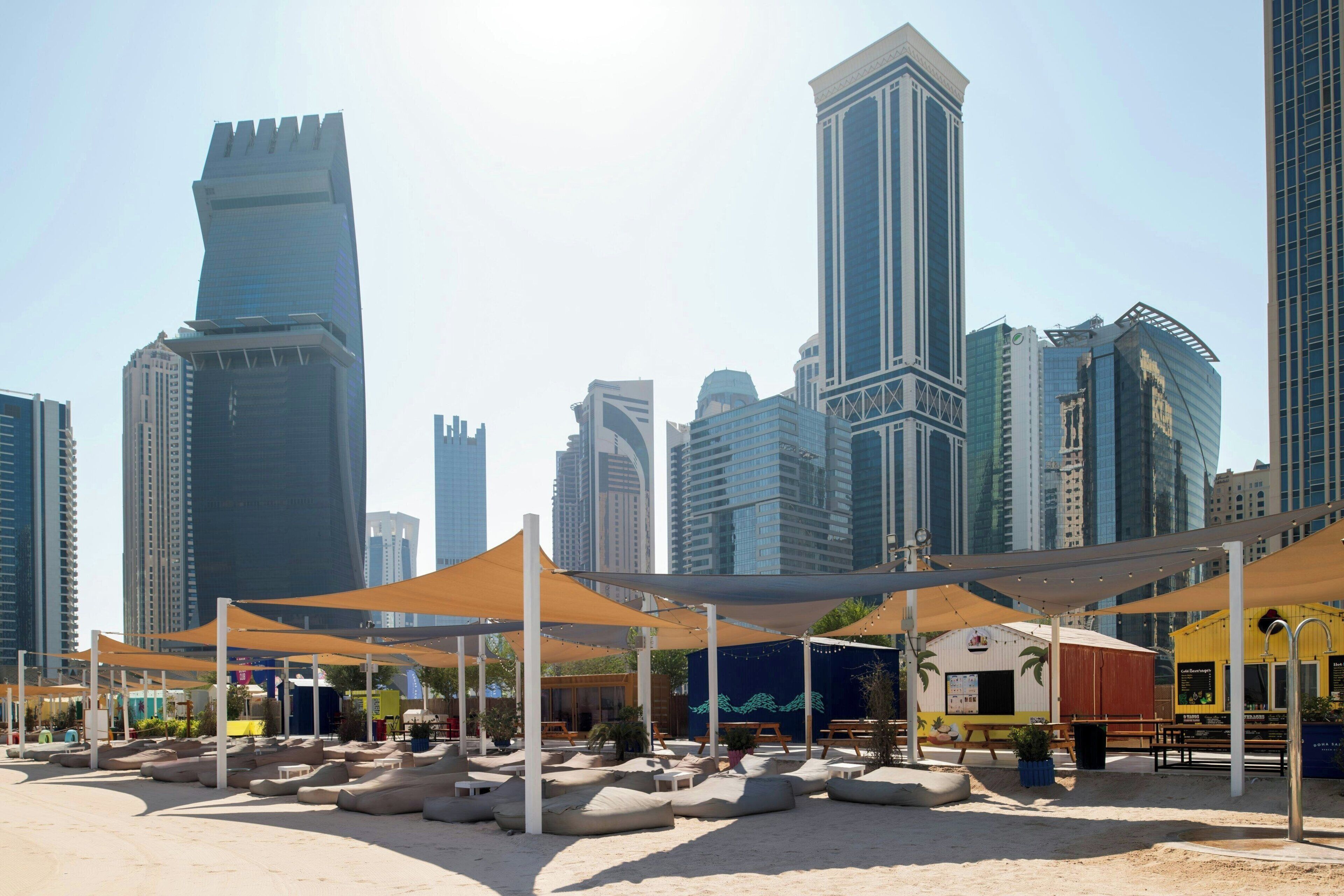 A modern urban setting with high-rise buildings, outdoor seating areas with cushions, and a small structure with a yellow facade.