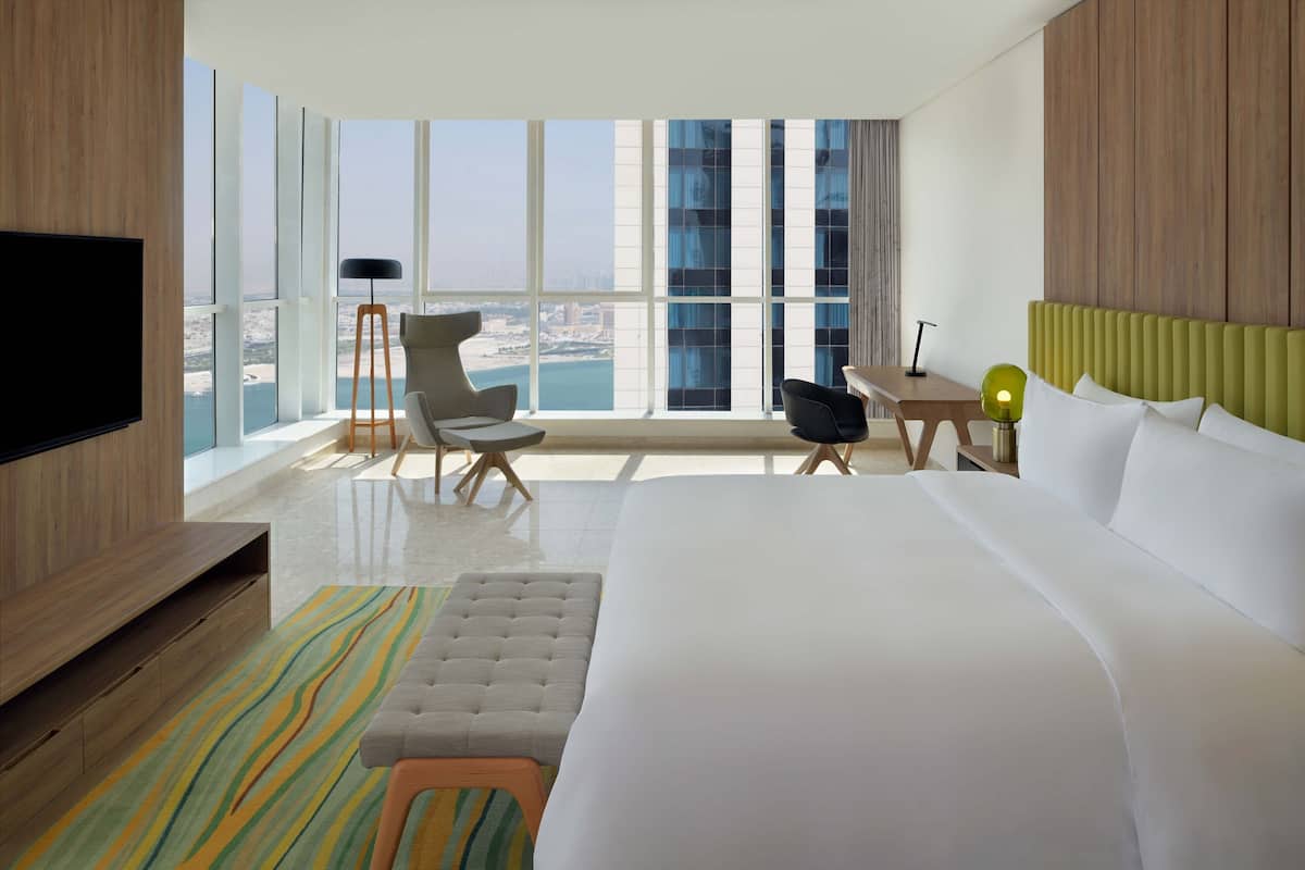 Habitación de hotel moderna con una cama grande, televisión, escritorio y una silla. La habitación tiene ventanas amplias que ofrecen vistas de la ciudad.