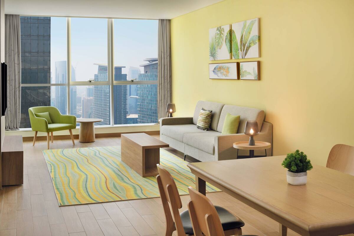 Una sala de estar moderna con sofá, sillas, una mesa de comedor y vistas a la ciudad a través de grandes ventanales.