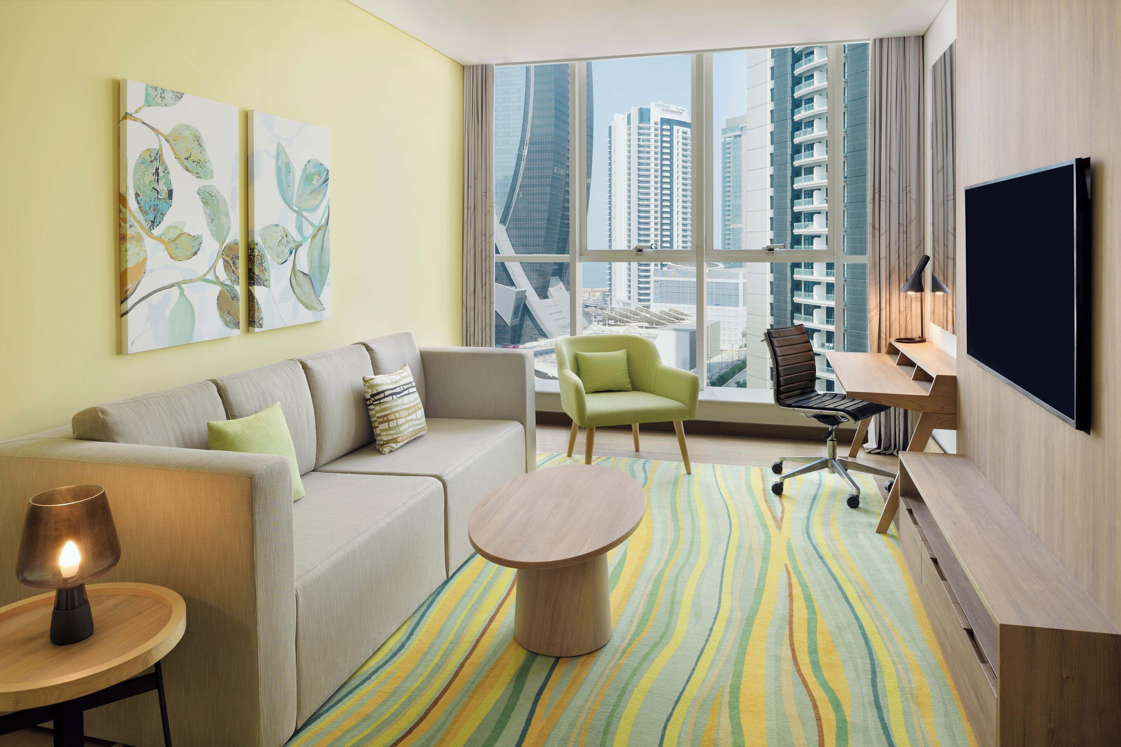 A modern hotel room with a sofa, armchair, desk, and large windows offering a city view.