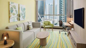 A modern hotel room with a sofa, armchair, desk, and large windows offering a city view.