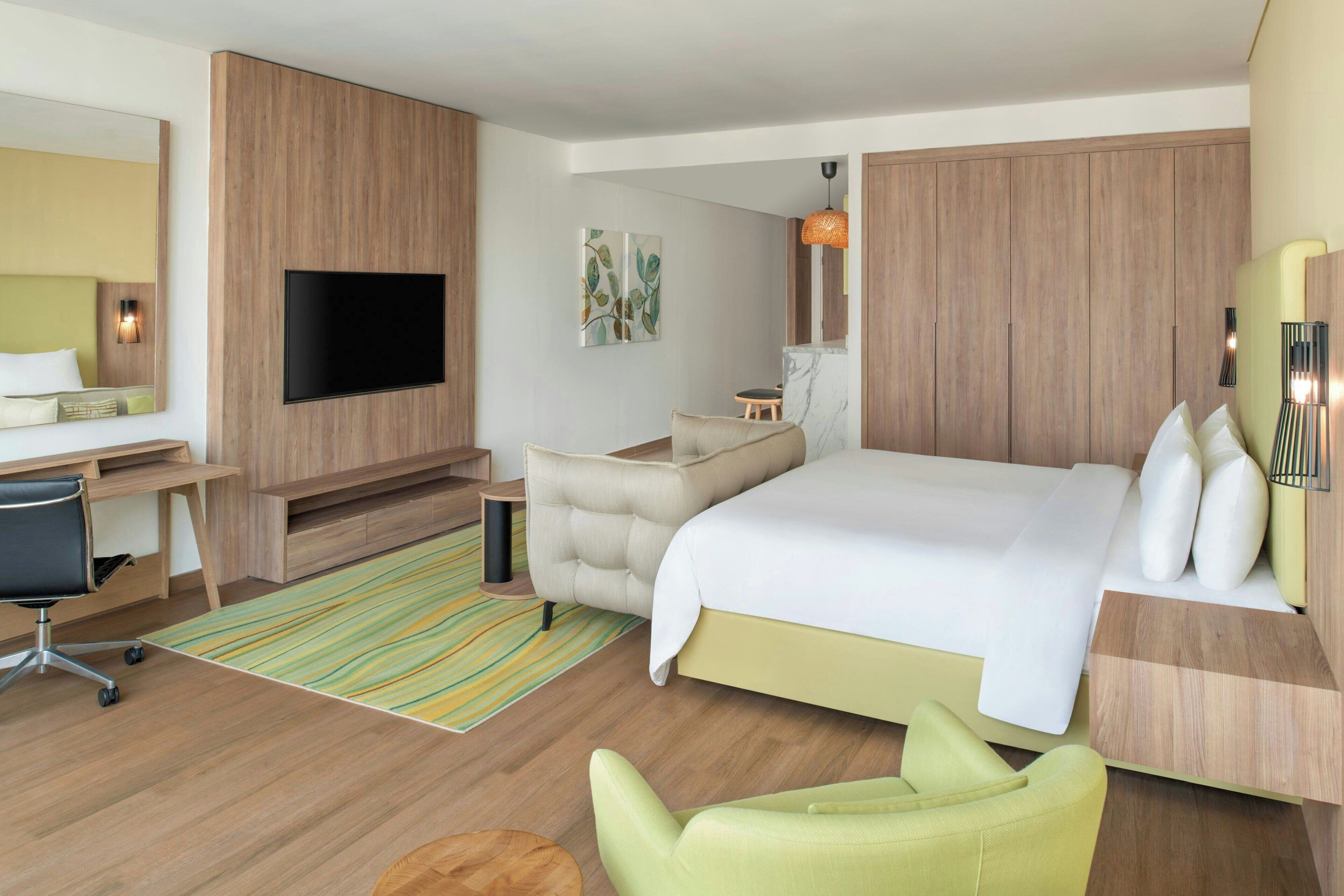 A modern hotel room with a large bed, a desk, a chair, a TV, and a small table.
