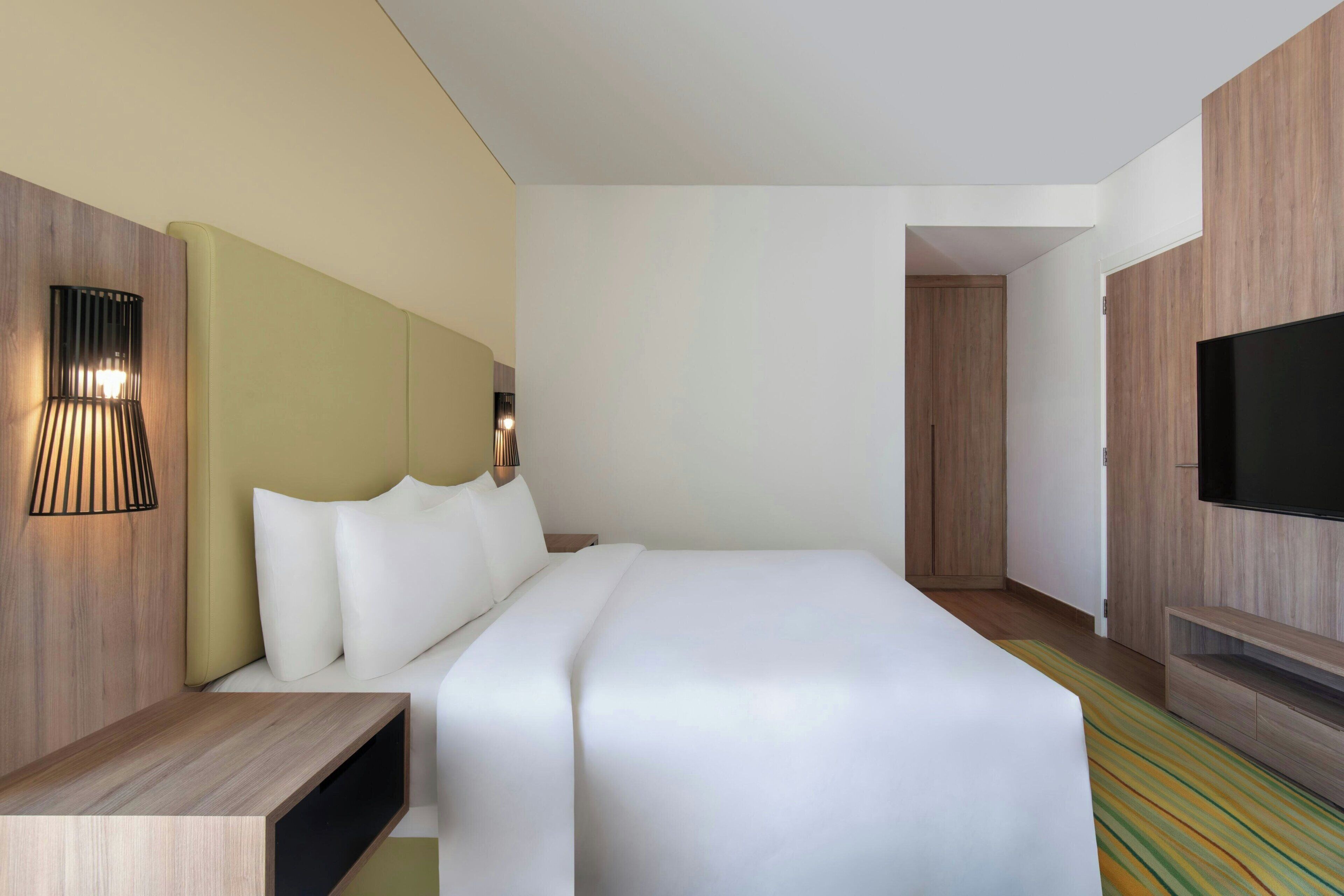 A modern hotel room with a large bed, a TV, a bedside table, and a wall-mounted lamp.