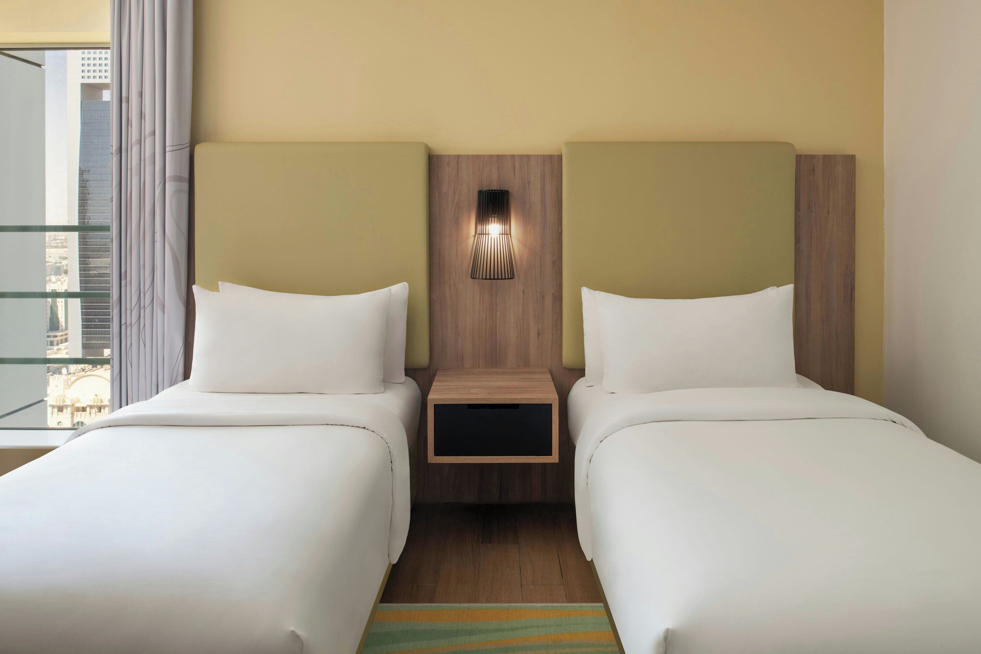 A hotel room with two beds, a nightstand with a lamp, and a view of a cityscape through a window.
