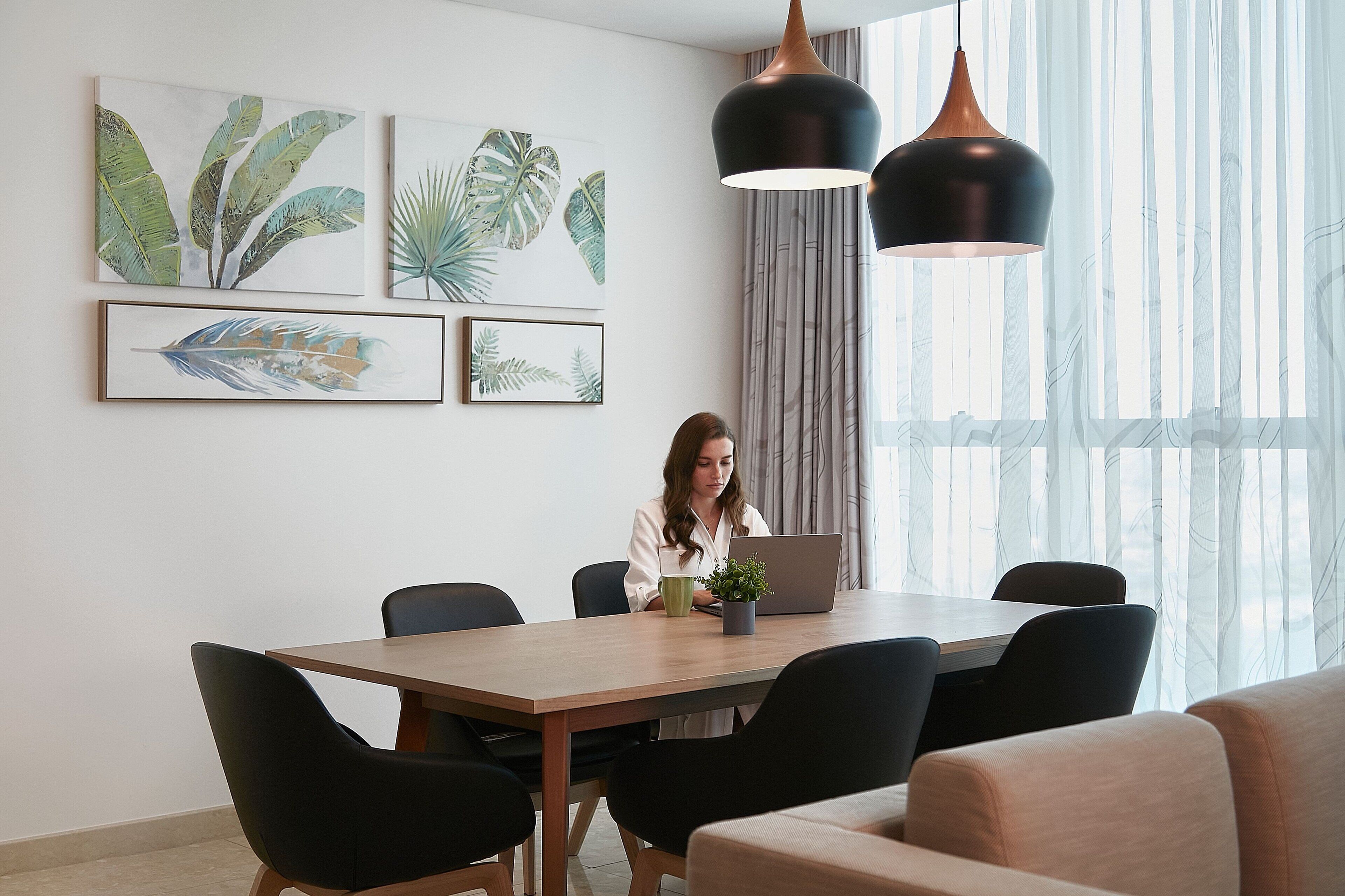 Un comedor moderno con una mesa de madera, sillas negras y un ordenador portátil. Hay láminas botánicas enmarcadas en la pared y una planta en maceta sobre la mesa.