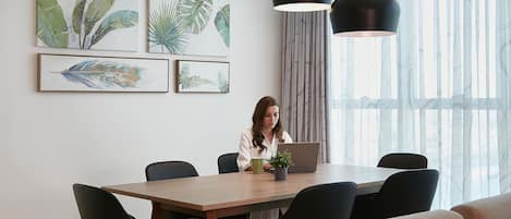 A modern dining area with a wooden table, black chairs, and a laptop. There are framed botanical prints on the wall and a potted plant on the table.