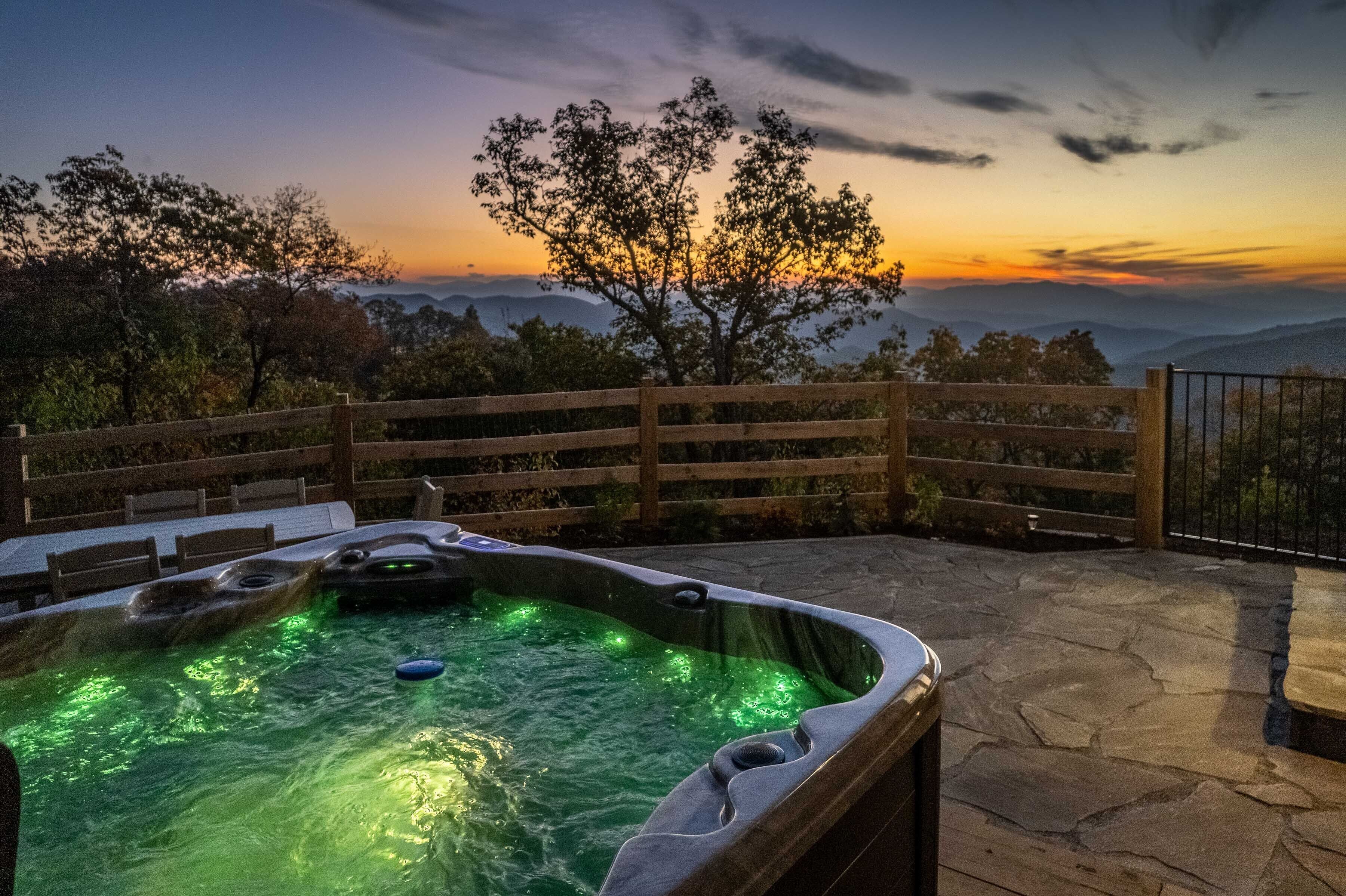 Un bain à remous avec une terrasse en pierre et une clôture en bois, offrant une vue sur un paysage montagneux au coucher du soleil.