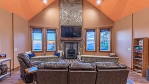 Un salon chaleureux avec une cheminée en pierre, des fauteuils en cuir et de grandes fenêtres donnant sur l’extérieur.