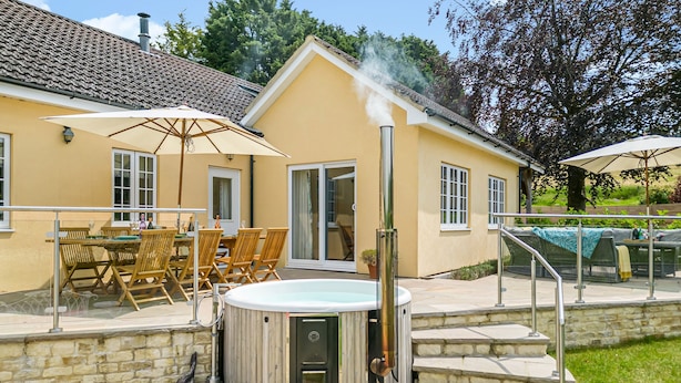 A hot tub in front of a house with a patio area, outdoor furniture, and a stone wall. - The Anchorage @ Nables Farm (Chippenham)