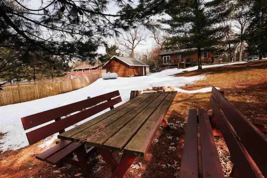 A wooden bench in a snowy outdoor area with a house in the background.