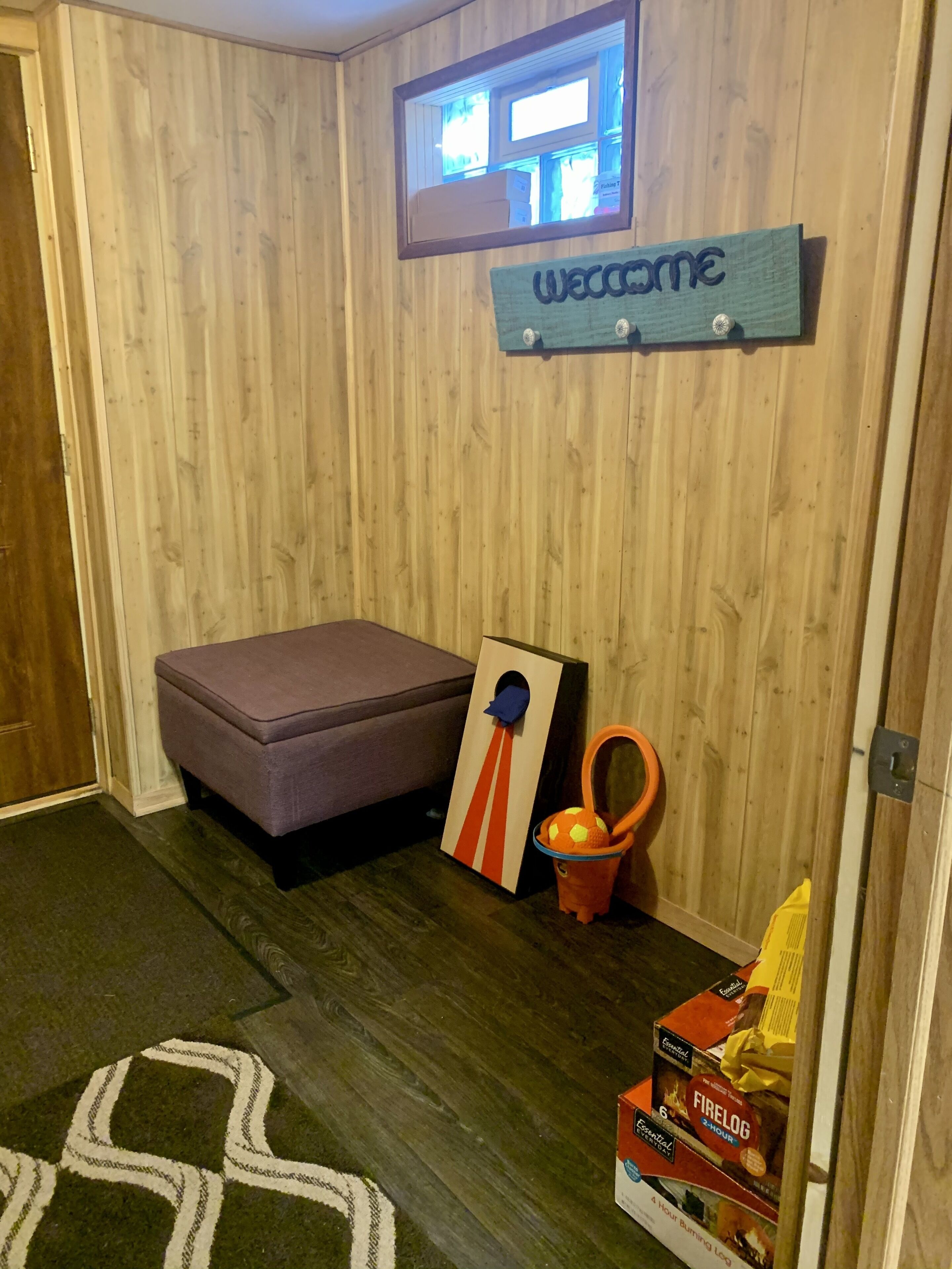 Ein Zimmer mit einer holzverkleideten Wand, einem Fernseher über einem 'Welcome'-Schild, einer violetten Bank, einem kleinen orangen Spielzeug und einem Kaminholzfach am Boden.