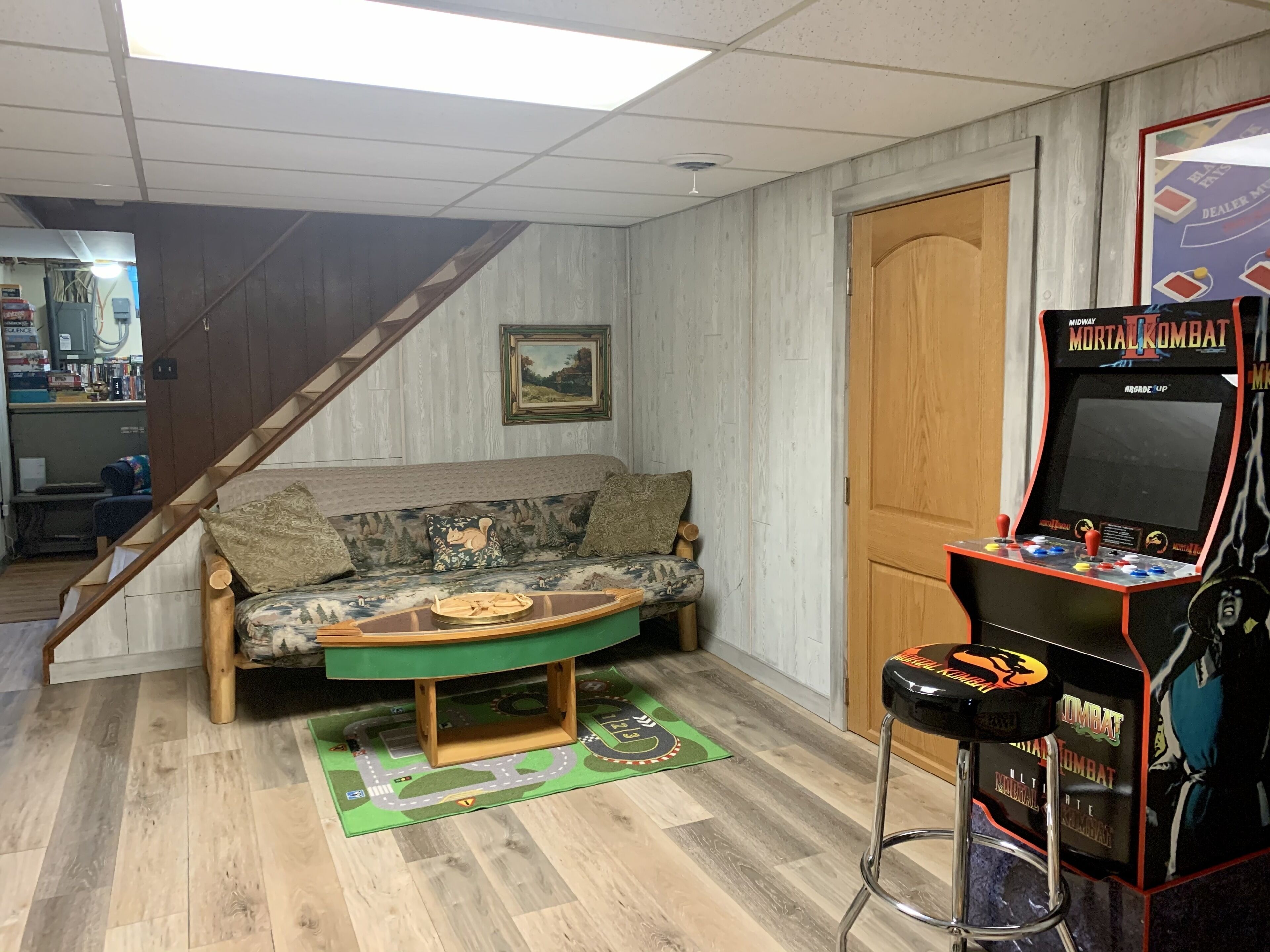 Una habitación con una escalera de madera, un sofá con cojines estampados, un tapete verde y una máquina recreativa retro.