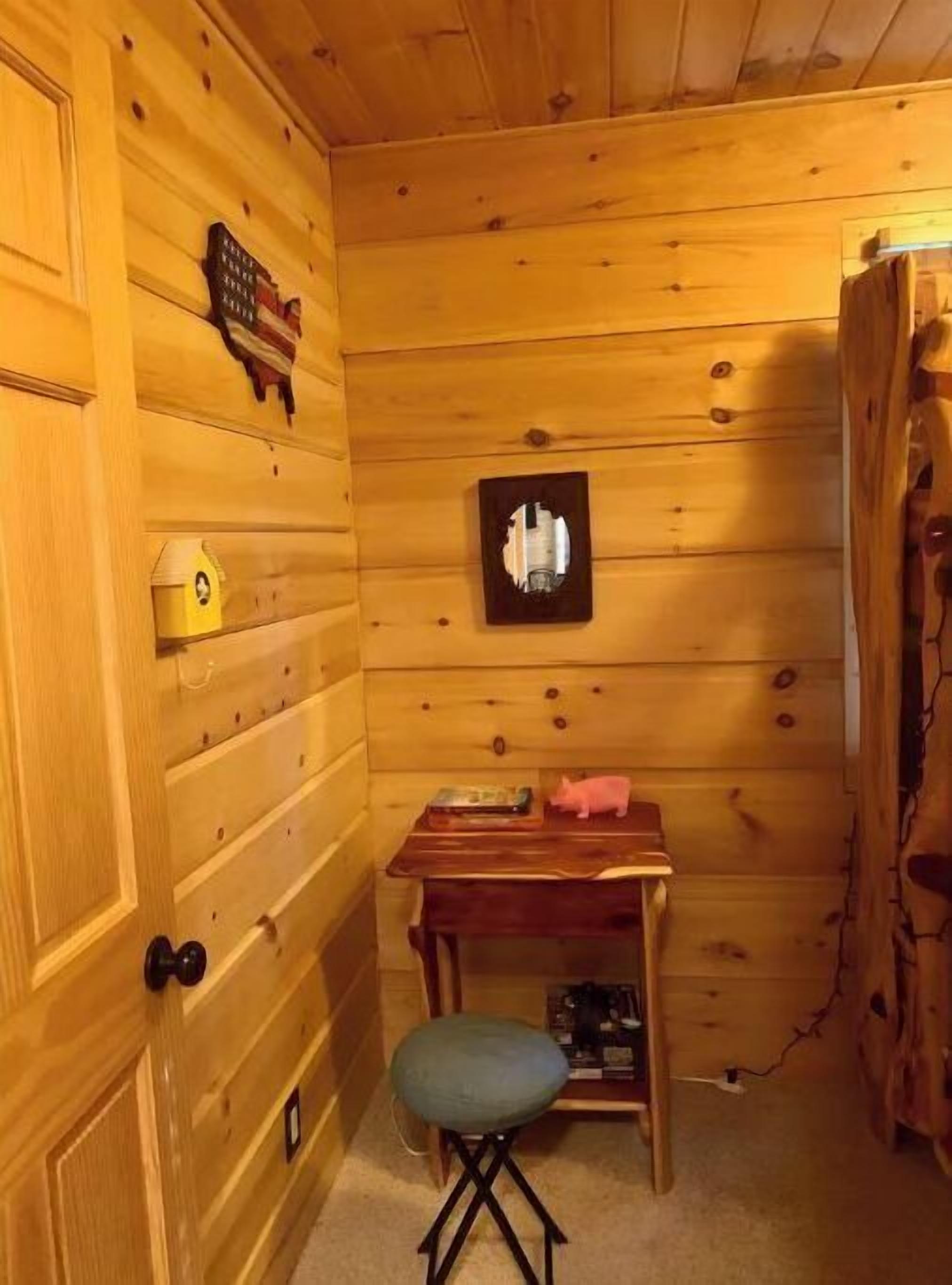 A wooden cabin interior with a wooden door, a small table with a book and a piggy bank, a green cushioned stool, and a framed picture on the wall.