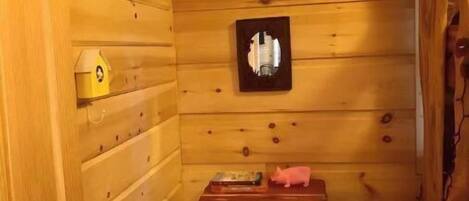 A wooden cabin interior with a wooden door, a small table with a book and a piggy bank, a green cushioned stool, and a framed picture on the wall.