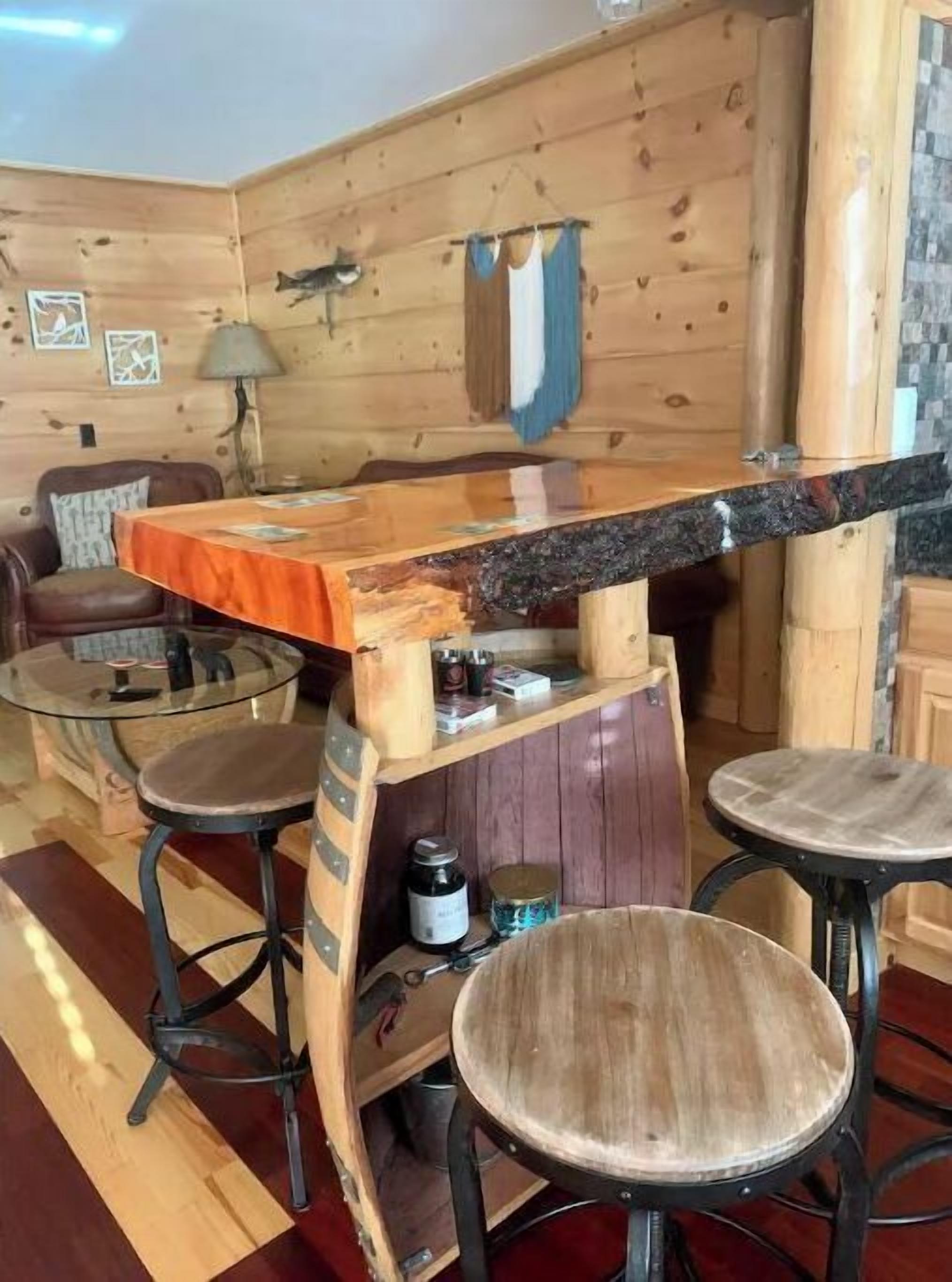 A rustic wooden interior with a bar area, two stools, a table, and a lamp.