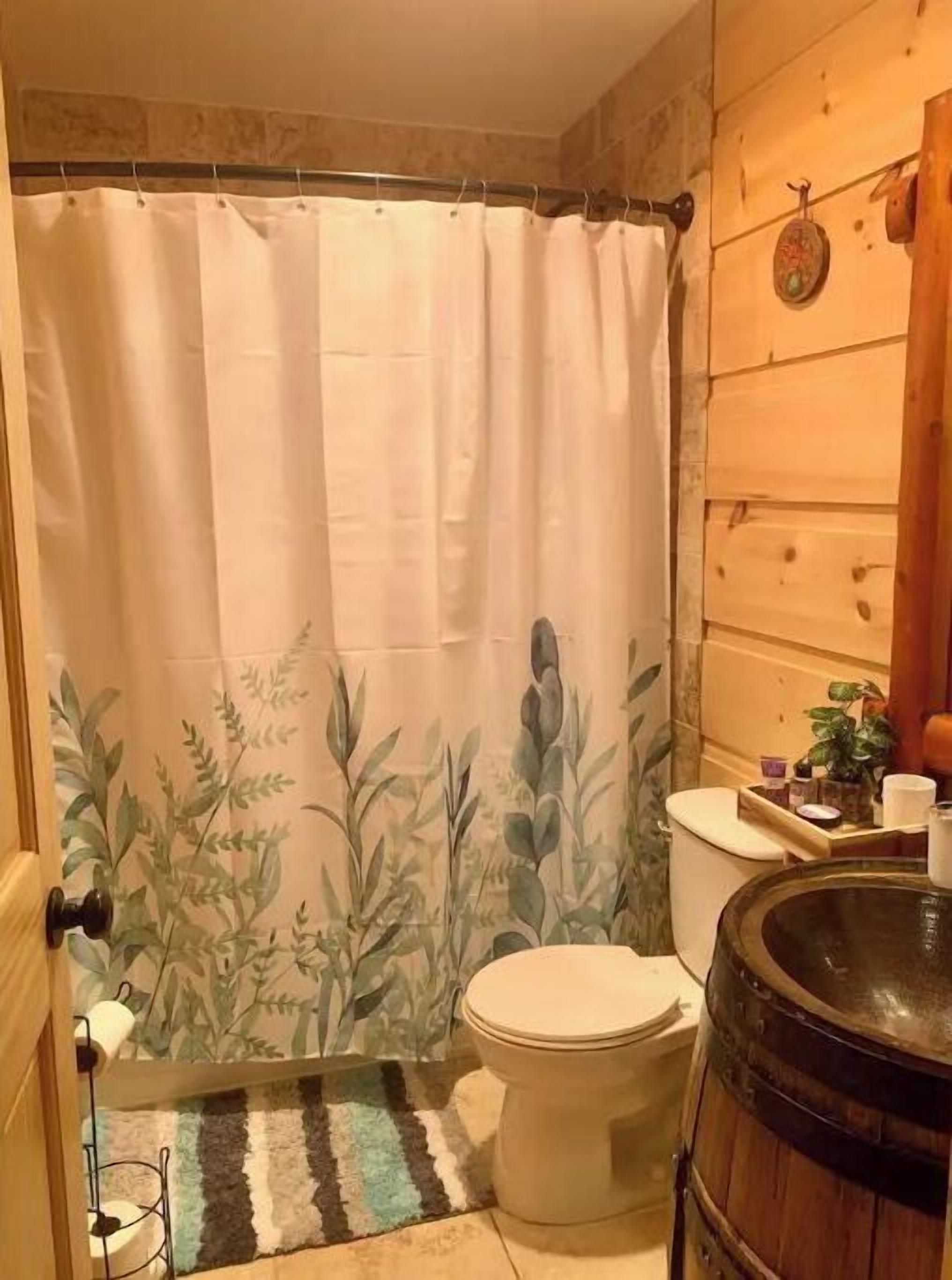 Een badkamer met een douchegordijn met een bladerenpatroon, een toilet, een vloerkleed en een houten vat.