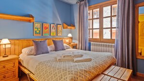 A bedroom with a wooden bed, a nightstand, a bench, a window with curtains, and wall art. - Luderna - Apartamento D2 Suriguera de Canillo (Canillo)
