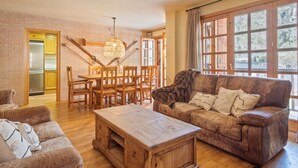 A cozy living room with a wooden coffee table, a brown sofa with cushions, a dining area with wooden chairs and a table, and large windows with curtains. - Luderna - Apartamento D2 Suriguera de Canillo (Canillo)