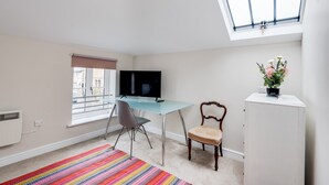 Una habitación pequeña y luminosa con escritorio, silla y televisor. Cuenta con una ventana con persianas, un radiador y una alfombra colorida.