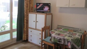 Una habitación con televisión sobre un mueble bajo, un armario, una mesa con mantel, una silla y una ventana con cortinas.
