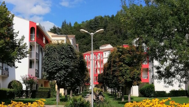 Un sendero conduce a un edificio de fachada roja y ventanas arqueadas, rodeado de follaje y parterres de flores.