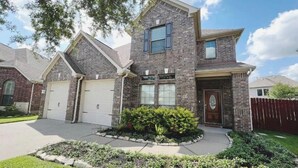 A two-story house with a stone facade, two garage doors, and a landscaped front yard with a variety of plants. - Spacious | Large Groups | Katy Mall, Parks! (Katy)