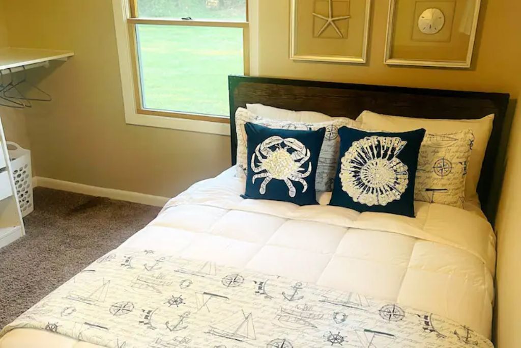 Una cama bien hecha con cubrecama de temática náutica y almohadones decorativos, una ventana con vista a un césped verde y un reloj montado en la pared.