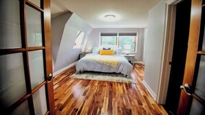 Un dormitorio con piso de madera, una cama con cubrecama blanco y amarillo, una mesita de noche y una ventana con vistas al exterior.