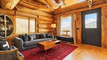 A log cabin interior with a sofa, coffee table, and a large window offering a mountain view.