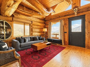 A log cabin interior with a sofa, coffee table, and a large window offering a mountain view.