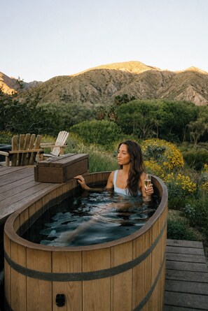 A wooden hot tub with a person inside, surrounded by a deck and natural landscape.