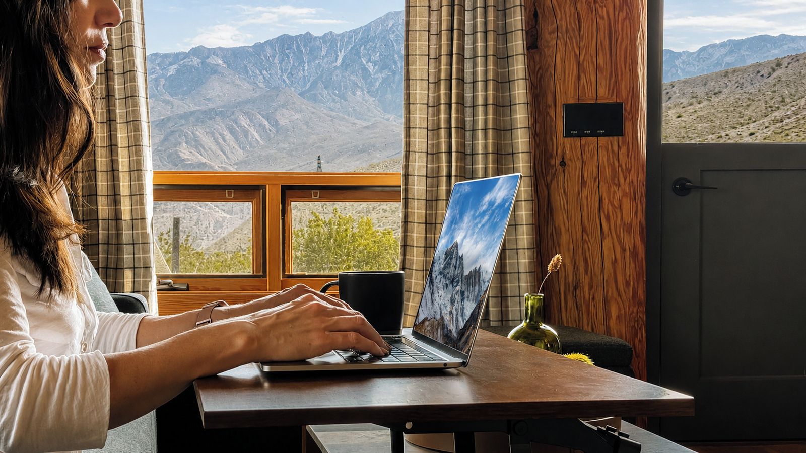 Une personne utilise un ordinateur portable à une table en bois, avec vue sur une montagne par la fenêtre.