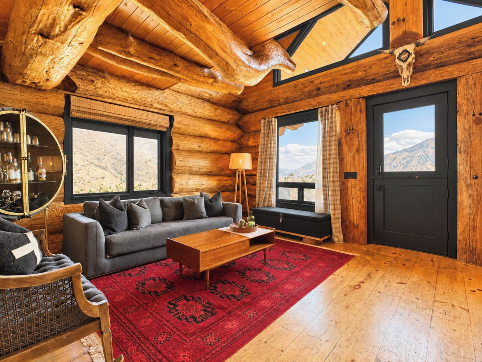Interior de uma cabana de tronco com sofá, mesa de centro e vista para as montanhas através de grandes janelas.
